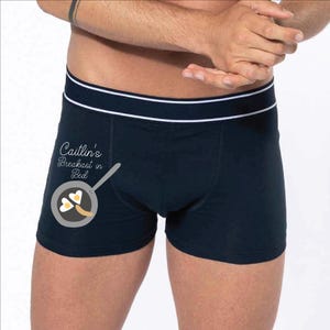 May include: Navy blue boxer briefs with a white elastic waistband featuring two thin horizontal stripes. The right leg displays a graphic of a frying pan containing two heart-shaped fried eggs and the text "Caitlin's Breakfast in Bed".