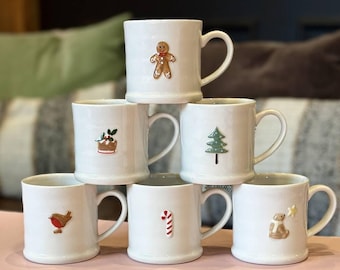 May include: A stack of seven white ceramic mugs with Christmas-themed designs. Each mug features a different embossed design, including a gingerbread man, Christmas tree, candy cane, and a robin. The mugs are arranged on a pink surface.