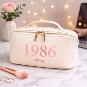 May include: A cream-colored cosmetic bag with a gold zipper and handle. The bag has the year "1986" and the name "Olivia" printed in pink. A makeup brush and two rings are in the foreground. A vase with a pink rose is in the background.