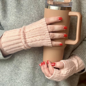 May include: A pair of light pink cable knit fingerless gloves. The gloves are worn by a person holding a tan insulated tumbler with a handle. The person is wearing a gray sweater. The person's nails are painted red.