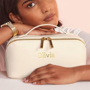 May include: A white leather cosmetic bag with a gold zipper and the name "Olivia" embossed in gold letters.