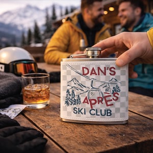 May include: A stainless steel hip flask with a white and grey chequered design. The flask has the text "DAN'S APRES SKI CLUB" in red and blue, with a mountain and ski lift graphic. A glass of amber liquid is nearby.