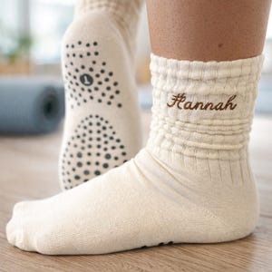 Può includere: Calzini color crema con polsino a coste e il nome "Hannah" ricamato in marrone. Le suole hanno punti antiscivolo neri. I calzini sono progettati per il comfort e l'aderenza, adatti per yoga o relax.