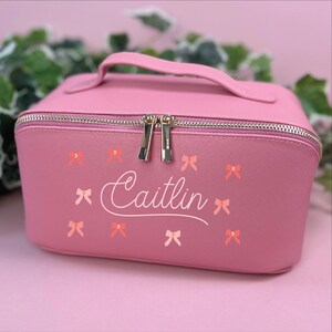 May include: A pink makeup bag with a handle and a zipper closure. The bag is decorated with small bow graphics and the name "Caitlin" in a cursive font. The bag is sitting on a pink surface.