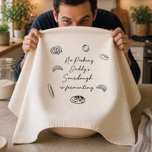 May include: A cream-colored sourdough bread cover with black illustrations of baked goods and the text "No Peeking Daddy's Sourdough is fermenting." The cover is held over a bowl by a person.