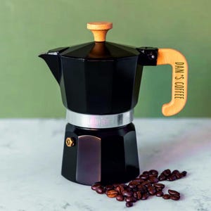 Op de afbeelding: Een zwarte espressomaker met een houten handvat en knop. Op het handvat staat "DAN'S COFFEE". Een stapel koffiebonen ligt naast de espressomaker. De espressomaker is ongeveer 20 cm hoog.