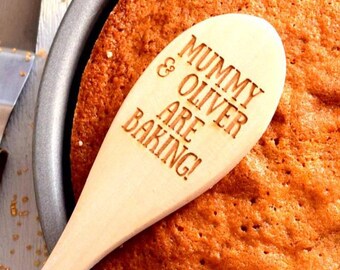 May include: A wooden spoon with the engraved text "MUMMY & OLIVER ARE BAKING!" rests on a freshly baked, round cake in a metal pan. The cake has a golden-brown crust. The spoon is light-colored.