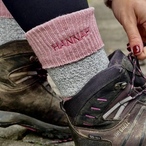 May include: A person wearing gray and pink knitted leg warmers with the name "HANNAH" embroidered on the pink cuff. The leg warmers are worn with brown hiking boots. The boots are being laced up.