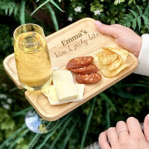 Könnte beinhalten: Ein Holzserviertablett mit dem Text "Emma's Wine & picky bits" enthält ein Glas Wein, Käse, Wurstscheiben und Kartoffelchips. Das Tablett wird von einer Person gehalten, mit einem grünen, blättrigen Hintergrund.