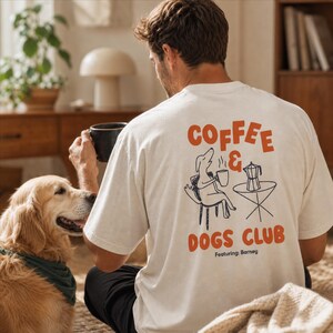 May include: Cream-coloured t-shirt with the words "COFFEE & DOGS CLUB" in orange, featuring a dog illustration. A golden retriever dog with a green bandana sits nearby. The shirt also says "Featuring: Barney".