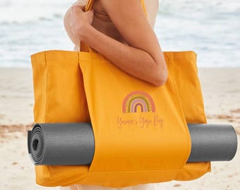 Op de afbeelding: Mosterdgele yogatas met een grijze yogamat. De tas heeft een regenboogafbeelding en de tekst "Yasmin's Yoga Bag". De tas wordt op het strand gedragen.