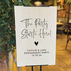 Op de afbeelding: Een houten bord met de tekst "The Party Starts Here!" in zwart script, een zwart hart en de namen "Caitlin & Jake" met "Engagement Party" en de datum "12.10.24". Het bord staat op een zwarte standaard.