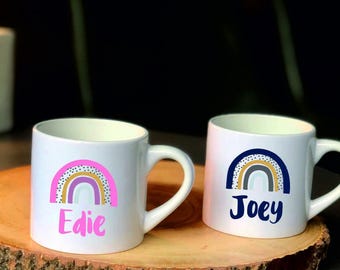 Op de afbeelding: Twee witte keramische mokken met regenboogontwerpen en namen. Eén mok heeft een roze regenboog met de naam "Edie" in roze. De andere mok heeft een blauwe, grijze en gele regenboog met de naam "Joey" in blauw.