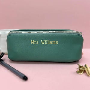 May include: A teal green pencil case with a silver zipper and the name "Mrs Williams" in gold lettering. The rectangular case is made of a textured material and is sitting on a pink surface.