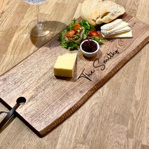May include: A wooden serving board with the text "The Smiths" engraved on it. The board holds cheese, bread, salad, and a small dish of jam. A glass of water sits to the left of the board. A leather strap is attached to the board.