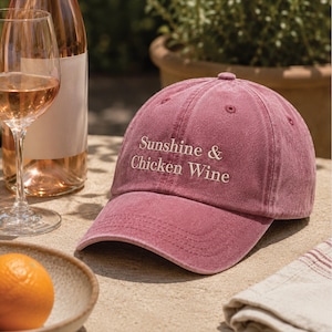 May include: A dusty rose baseball cap featuring the embroidered text Sunshine & Chicken Wine in a light cream colour. The cap has a structured crown, a curved brim, and stitched eyelets, presenting a classic six-panel design with a vintage finish.
