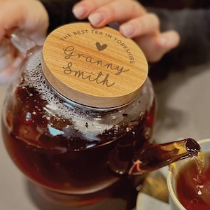 Puede incluir: Una tetera de vidrio llena de té, con una tapa de madera que dice "THE BEST TEA IN YORKSHIRE" y "Granny Smith". El té se está vertiendo en una taza de té. La tetera tiene un pico curvo y un cuerpo redondo.