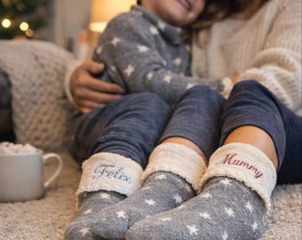 May include: Grey star-patterned socks with cream cuffs, one embroidered with "Felix" and the other with "Mummy". A child and an adult are wearing the socks. A mug of hot chocolate is visible in the background.