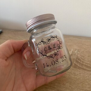 May include: A small, clear glass jar with a handle and a rose gold lid. The jar features a heart-shaped design with the initials "C & L" and the date "14.08.23". The design is in black. The jar is likely for a wedding or special event.