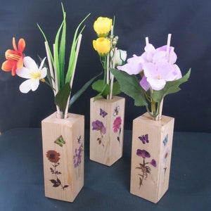 May include: Three wooden vases with different floral designs and dried flowers. Each vase has a different colour scheme: one with yellow flowers, one with purple flowers, and one with orange and white flowers.