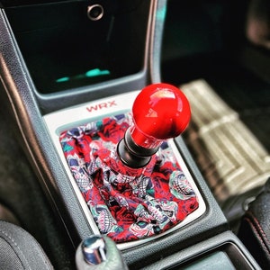 May include: A red and black car interior with a red gear shift knob and a custom skull and rose patterned gear shift boot. The boot is surrounded by a silver trim and the word "WRX" is visible on the console.