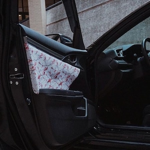 Pode incluir: Um carro preto com a porta do lado do motorista aberta, revelando um tecido estampado em branco e rosa que cobre o painel da porta. O interior do carro é preto e o volante é visível.