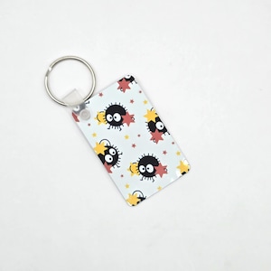 May include: A rectangular keyring with a repeating pattern of black, round characters with large eyes and red and yellow star accents on a light blue background. The keyring has a silver metal ring.