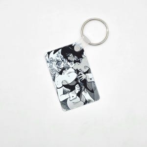May include: A rectangular keychain featuring a black and white manga-style illustration of two figures in intimate embrace. The keychain has a clear plastic casing and a silver metal keyring.