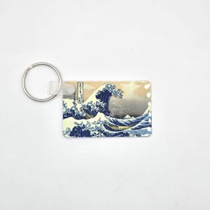 May include: A rectangular keychain featuring the iconic Japanese artwork "The Great Wave off Kanagawa." The design showcases a large wave in shades of blue and white, with a light beige sky. A silver key ring is attached.