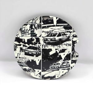 May include: Black and white round coaster with a manga-style design featuring a car racing scene. The coaster has a cut-out design on the side.