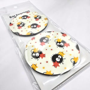 May include: Two round car coasters with a white background and a repeating pattern of black and white cartoon characters with red stars. The text "Car Coasters" is printed on the packaging.