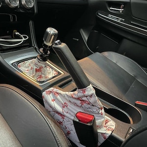 Könnte beinhalten: Ein weißer und roter, floral gemusterter Schaltknauf-Stiefel mit einem silbernen Schaltknauf. Der Stiefel ist in der Mittelkonsole eines Autos installiert.