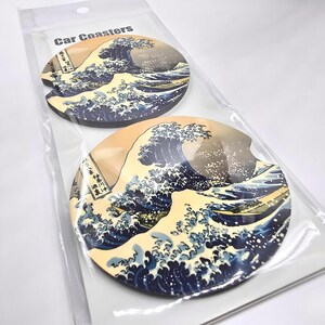 May include: Two round car coasters featuring a Japanese woodblock print of "The Great Wave off Kanagawa" by Katsushika Hokusai. The coasters are white with blue and yellow accents. The text "Car Coasters" is printed on the packaging.