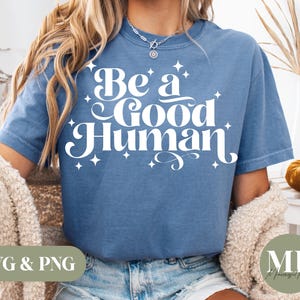 May include: A blue t-shirt with the words "Be a Good Human" in white script. The text is surrounded by small starburst designs. The shirt is part of a clothing collection.
