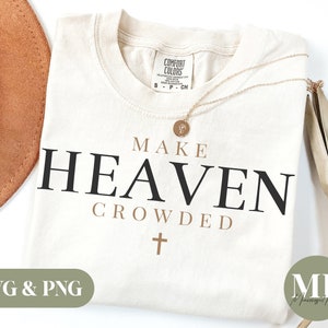 Pu&ograve; includere: Una maglietta bianca con il testo "MAKE HEAVEN CROWDED" in lettere nere e marroni. Una piccola croce si trova sotto il testo. La maglietta &egrave; su uno sfondo bianco con un cappello marrone e una pianta secca.