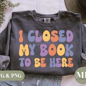 Op de afbeelding: Een grijs sweatshirt met de tekst "Ik heb mijn boek dicht gedaan om hier te zijn" in kleurrijke, afgeronde letters. De letters zijn in tinten oranje, geel, paars en blauw.