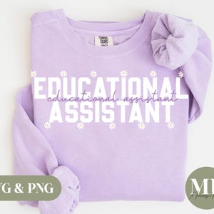 Puede incluir: Una sudadera de color lavanda con el texto "EDUCATIONAL ASSISTANT" en blanco con un patrón de margaritas alrededor del texto. El texto "educational assistant" está en una fuente más pequeña debajo del texto principal.