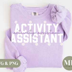 May include: A light purple sweatshirt with the words "ACTIVITY activity assistant ASSISTANT" in white block letters. Small white daisy graphics are interspersed. A scrunchie in the same color is on the right sleeve.