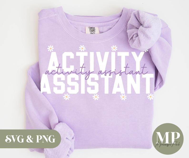 May include: A light purple sweatshirt with the words "ACTIVITY activity assistant ASSISTANT" in white block letters. Small white daisy graphics are interspersed. A scrunchie in the same color is on the right sleeve.