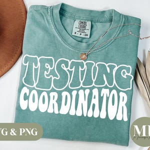 Peut inclure: Un t-shirt de couleur sarcelle avec l'inscription "TESTING COORDINATOR" en blanc, dans une police rétro. Un collier doré avec un pendentif est posé sur le t-shirt. Marque Comfort Colors.