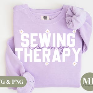 Puede incluir: Sudadera morada con el texto "Coser es mi terapia" en letras blancas con un diseño de margarita. El texto está en una fuente en negrita y de bloque.