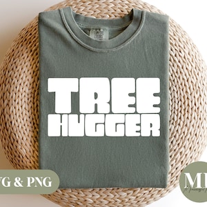 May include: A sage green t-shirt with the white text "TREE HUGGER" in a bold, blocky font. The shirt is displayed on a woven, light brown surface. The words "SVG & PNG" are in the lower left corner.