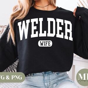 May include: Black sweatshirt with the word "WELDER" in large white block letters across the chest, with "WIFE" in a smaller white font underneath. The sweatshirt is worn by a person with blonde hair and blue jeans.
