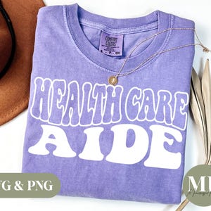Puede incluir: Camiseta morada con el texto "HEALTH CARE AIDE" en blanco, con letras de estilo retro. La camiseta está hecha de un material suave y cómodo.