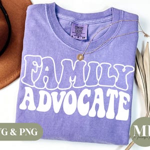 Puede incluir: Camiseta morada con el texto "FAMILY ADVOCATE" en blanco, con letras de estilo retro. La camiseta está hecha de un material suave y cómodo y tiene un aspecto desgastado.