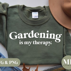 May include: A green sweatshirt with the text "Gardening is my therapy." in white lettering. The sweatshirt is laid flat on a surface with a brown hat and blue jeans.