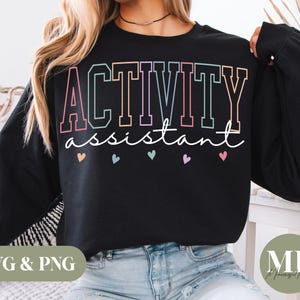 Op de afbeelding: Zwart sweatshirt met de woorden "ACTIVITY assistant" in kleurrijke blokletters. Onder de tekst staan kleine hartjes in verschillende pastelkleuren. Het sweatshirt wordt door een persoon gedragen.