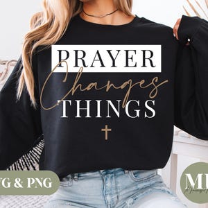 Peut inclure: Sweat-shirt noir avec l'inscription "PRAYER Changes THINGS" en blanc et or, avec une petite croix dorée en dessous. Le design est sur le thème religieux, adapté aux vêtements.