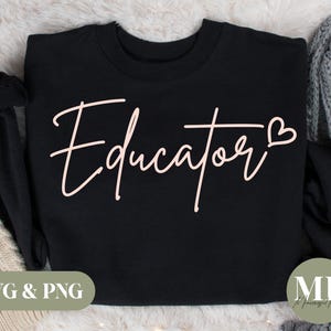 May include: Black sweatshirt with the word "Educator" in a cursive, light pink font. A small heart is next to the word. The bottom left corner has the text "SVG & PNG" in a green box. The sweatshirt is on a white, fluffy surface.