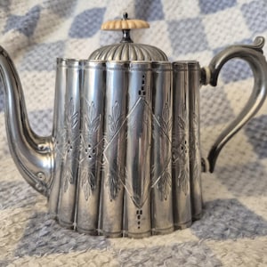 May include: A silver teapot with a fluted body and decorative leaf and diamond patterns. It features a curved spout and handle, and a lid with a decorative finial. The teapot is likely vintage or antique.
